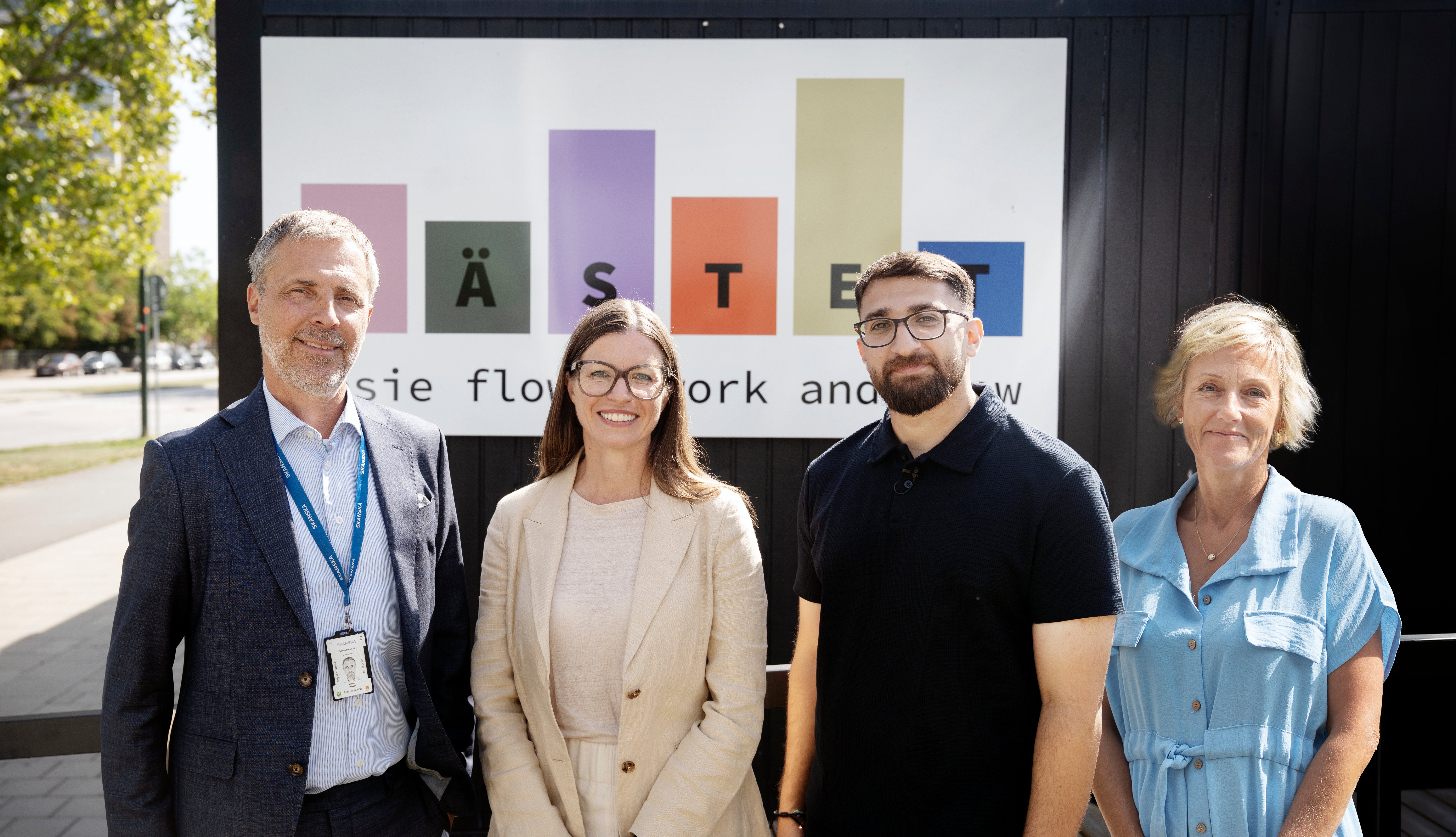 Fyra personer står framför en skylt med texten Nästet utomhus en solig dag. I bakgrunden syns en byggnad och grönska.