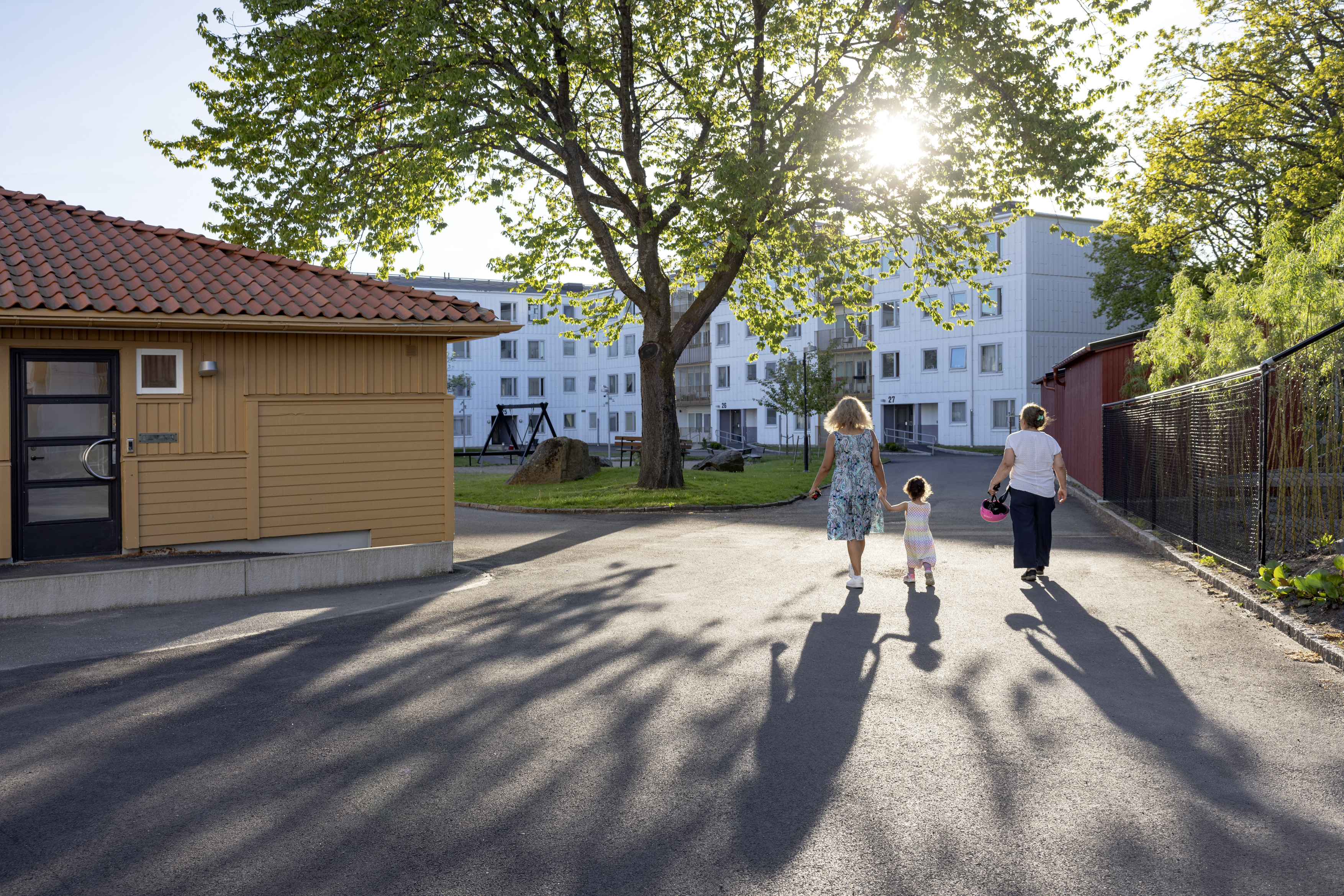 Tre personer går tillsammans genom ett soligt bostadsområde med flerfamiljshus, stora träd och en lekplats i bakgrunden. En vuxen håller ett barn i handen, och långa skuggor faller över gångvägen i kvällsljuset.