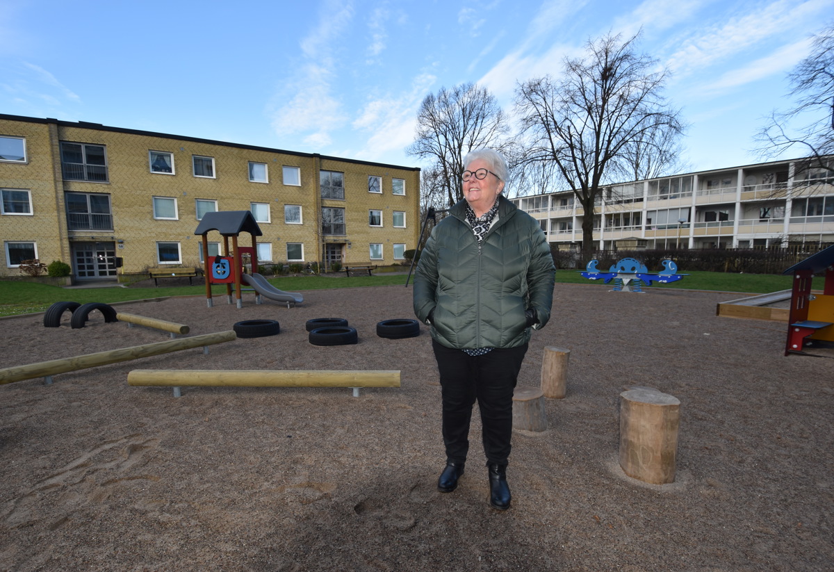 En person står på en lekplats omgiven av flerbostadshus. Det är en klar dag med blå himmel. Lekplatsen har gungor och klätterställningar, och träden i bakgrunden är utan löv. Personen bär en grön jacka och ser ut att njuta av den friska luften.