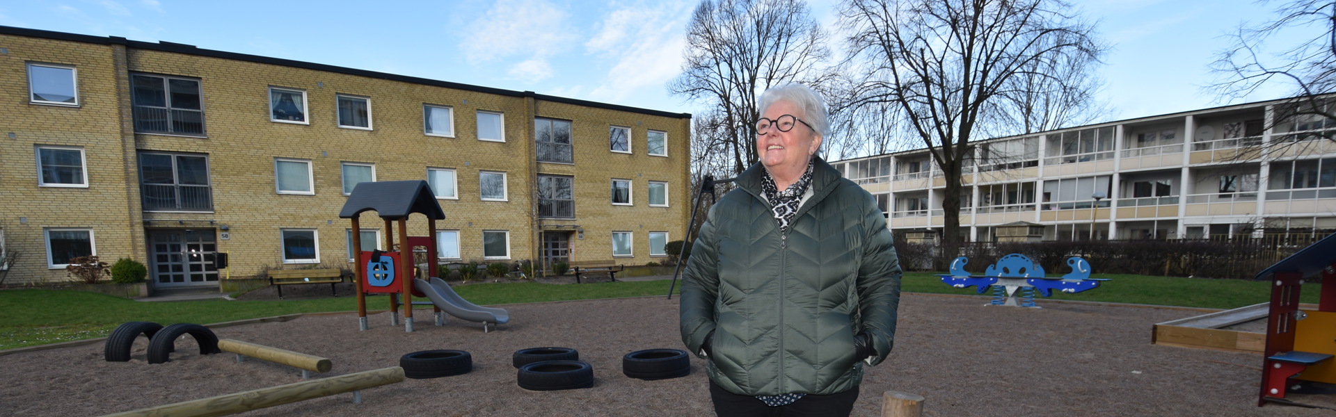 En person står på en lekplats omgiven av flerbostadshus. Det är en klar dag med blå himmel. Lekplatsen har gungor och klätterställningar, och träden i bakgrunden är utan löv. Personen bär en grön jacka och ser ut att njuta av den friska luften.
