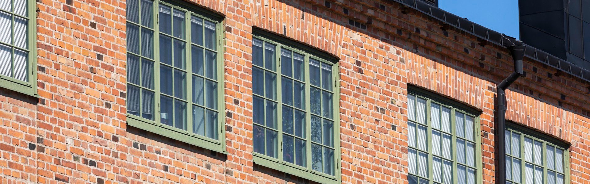 Övre delen av en tegelbyggnad med flera stora, grönramade fönster. Byggnaden har en traditionell röd tegelfasad och ett svart tak. Himlen är klar och blå.