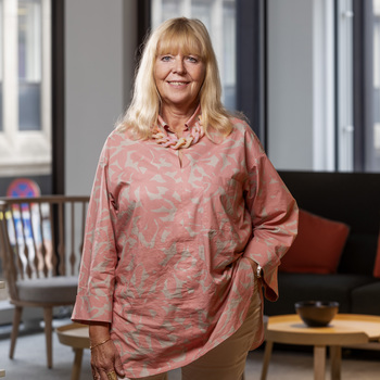 Agneta Kores. En kvinna med långt blont hår står inomhus och ler mot kameran. Hon bär en rosa mönstrad blus och ett halsband. Bakgrunden visar en modern inredning med stora fönster och en stol.
