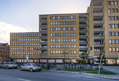 En bred bostadsbyggnad i gult tegel med stora fönster i regelbundet mönster. Husets bottenvåning rymmer butiker eller verksamheter med glasade entréer, och framför byggnaden syns parkerade bilar, cyklar och en liten grön yta med träd och gräs. Solen håller på att gå ner bakom huset och skapar ett mjukt ljus i himlen.