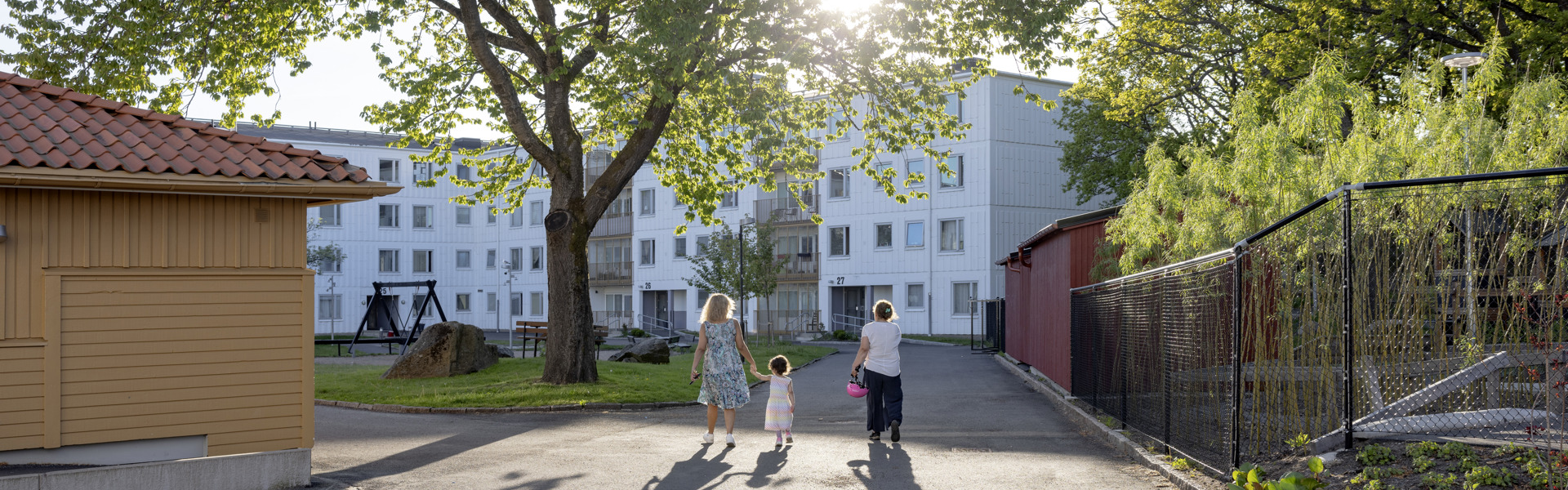 Bild från kvarter i Tynnered i Göteborg. Vita flerfamiljshus i bakgrunden. Solen lyser, en kvinna håller ett barn i handen och bredvid dem går en kvinna tillsammans med dem och håller en cykelhjälm i handen. Personerna ses bakifrån.