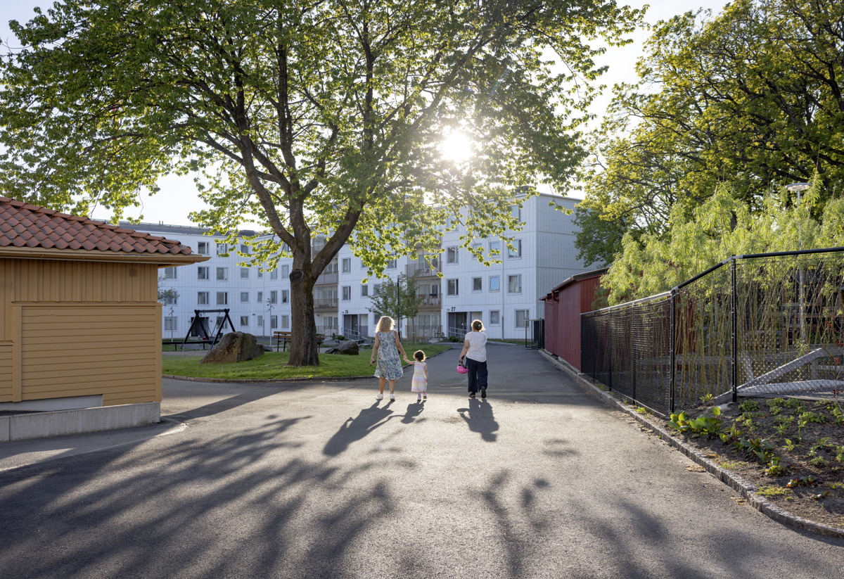 Bild från kvarter i Tynnered i Göteborg. Vita flerfamiljshus i bakgrunden. Solen lyser, en kvinna håller ett barn i handen och bredvid dem går en kvinna tillsammans med dem och håller en cykelhjälm i handen. Personerna ses bakifrån.