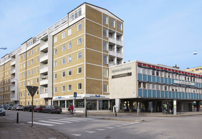 Ett hörn av ett bostadsområde med flerfamiljshus i tegel och en byggnad med blå och vita detaljer, under en klarblå himmel.