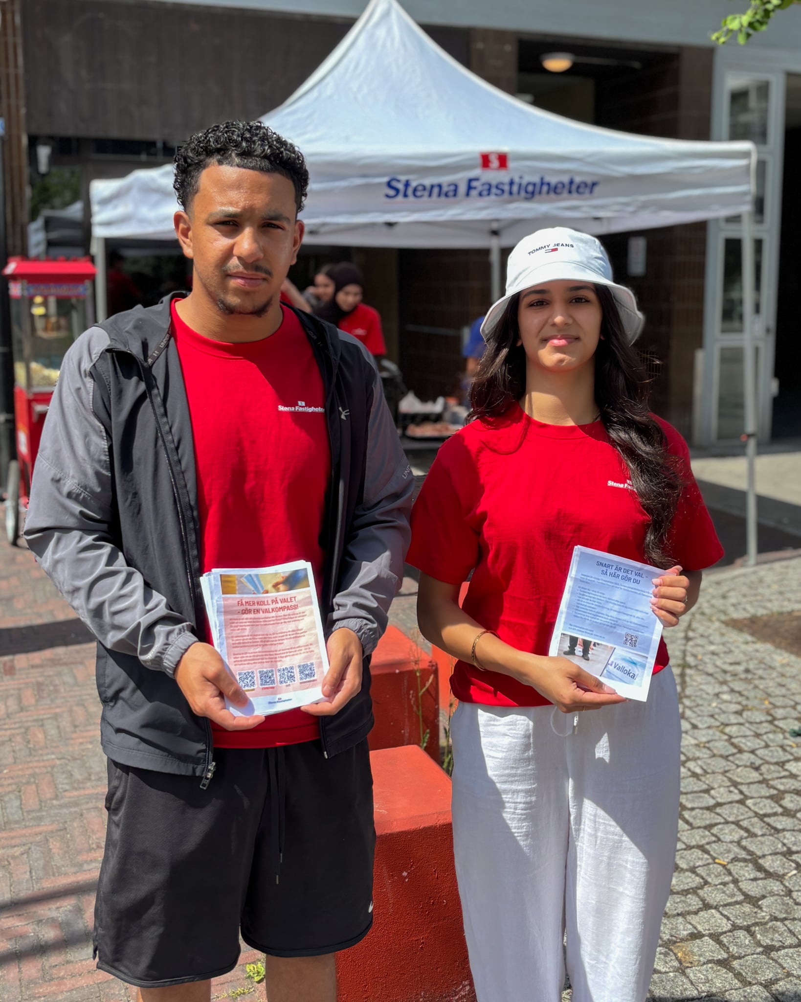 Två unga personer står framför ett tält med Stena Fastigheter-logotyp. De bär röda tröjor och håller i informationsblad med QR-koder. Bilden är tagen utomhus i solsken, på en plats med stenläggning och byggnader i bakgrunden.