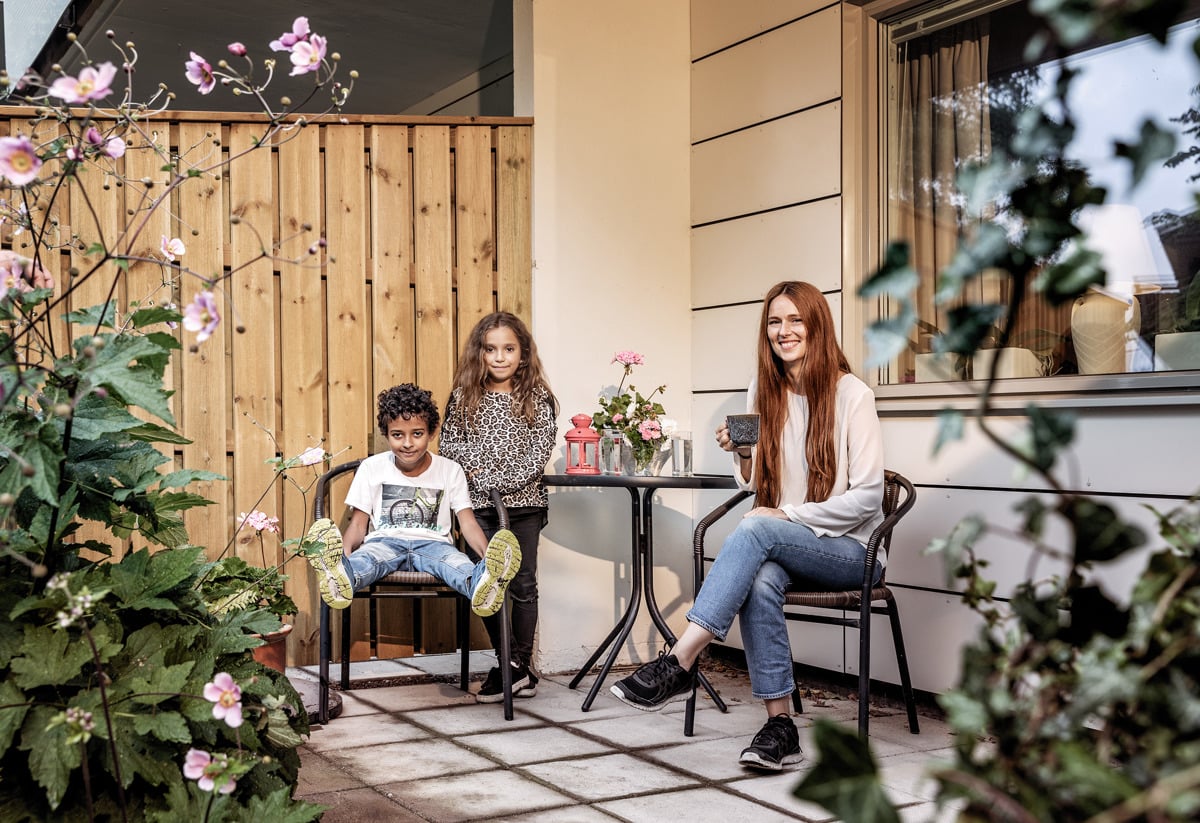 En leende kvinna och två barn sitter på en uteplats omgivna av blommor