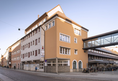 Gul och vit hörnbyggnad i stadsmiljö med flera våningar och en gångbro i glas som sträcker sig över gatan till en annan byggnad. Det står cyklar längs trottoaren, och några personer går längs gatan.