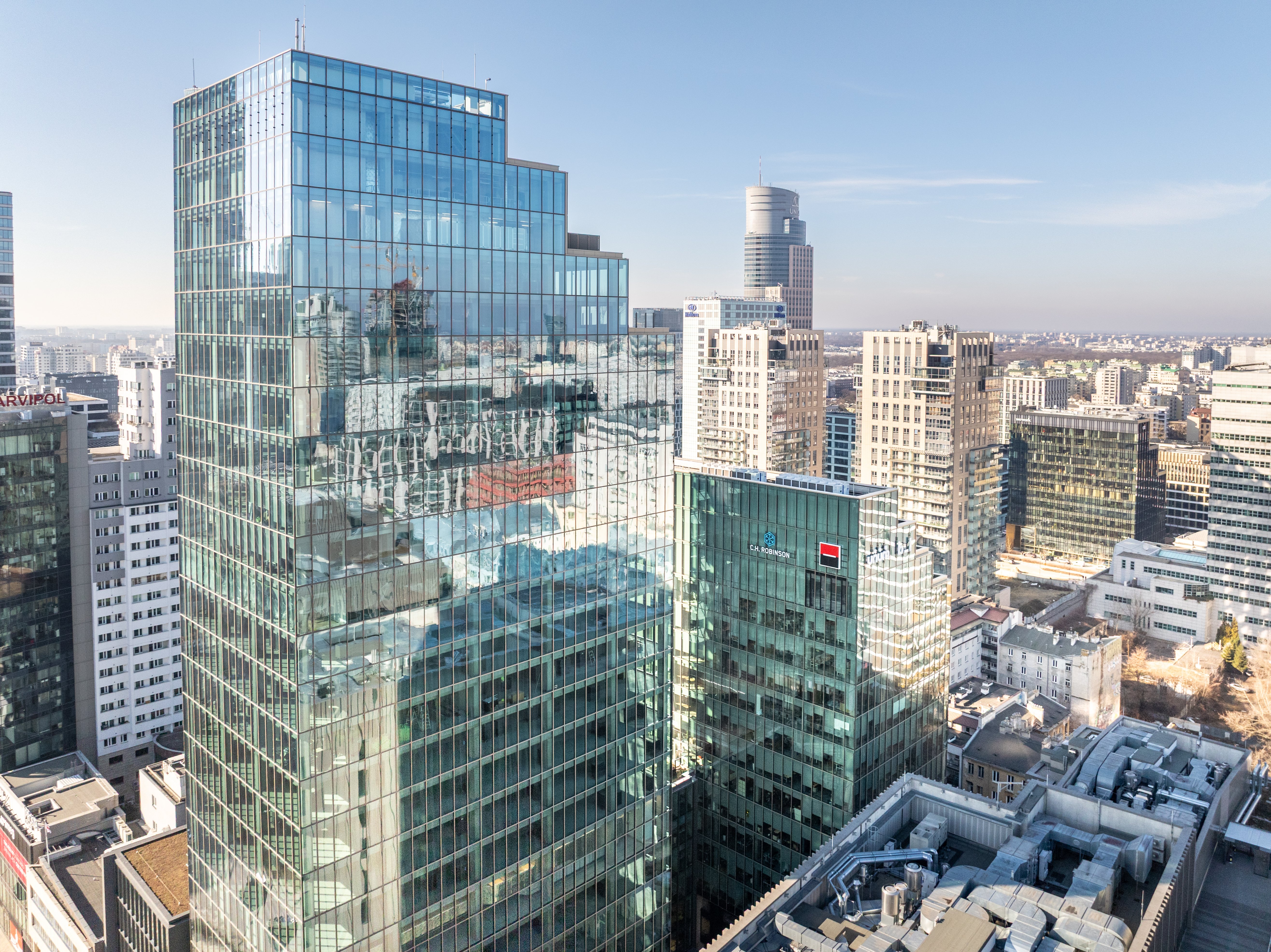 A modern cityscape with tall glass buildings on the left side of the image and a wide railway corridor running through the city to the right, surrounded by lower houses and office buildings in clear, sunny weather. Warsaw, Poland