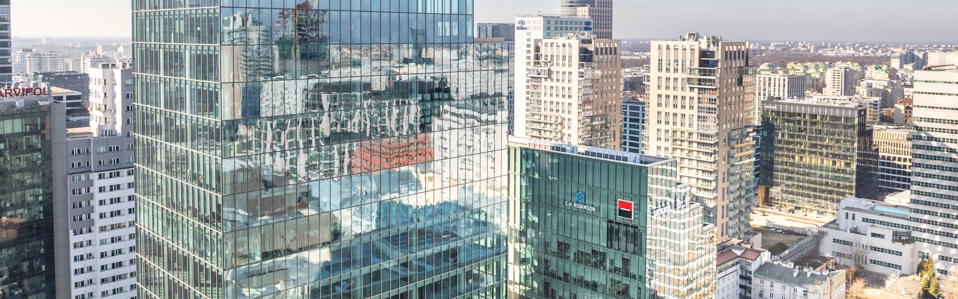 En modern stadsbild med höga glasbyggnader till vänster i bild och en bred järnvägskorridor som löper genom staden åt höger, omgiven av lägre hus och kontorsbyggnader i klart, soligt väder. Warszawa, Polen