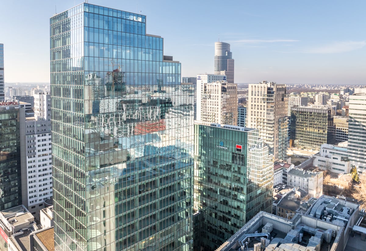 En modern stadsbild med höga glasbyggnader till vänster i bild och en bred järnvägskorridor som löper genom staden åt höger, omgiven av lägre hus och kontorsbyggnader i klart, soligt väder. Warszawa, Polen