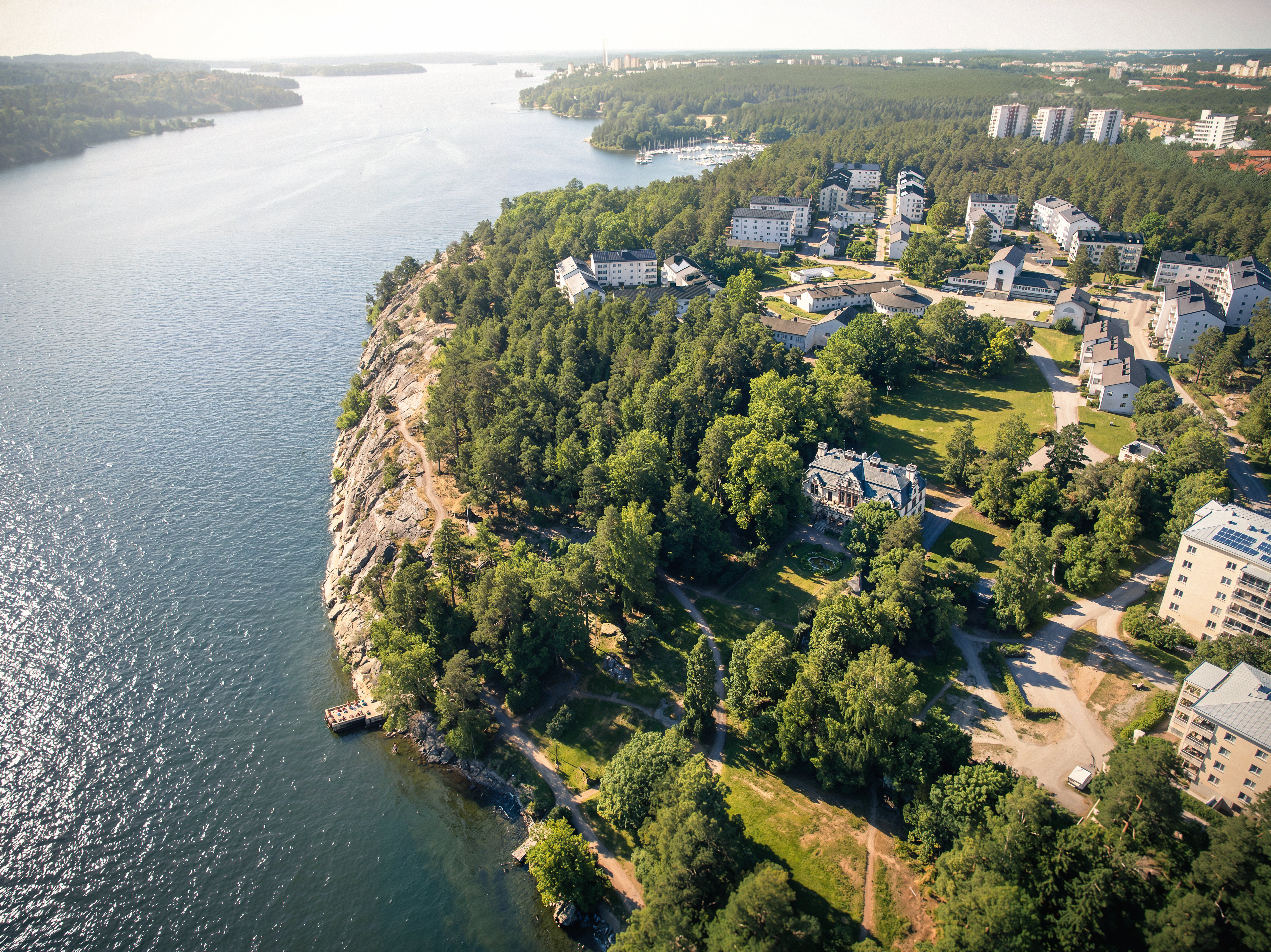 Flygfoto över ett bostadsområde vid vatten, med flerbostadshus, grönskande skogspartier och klippor längs strandlinjen.