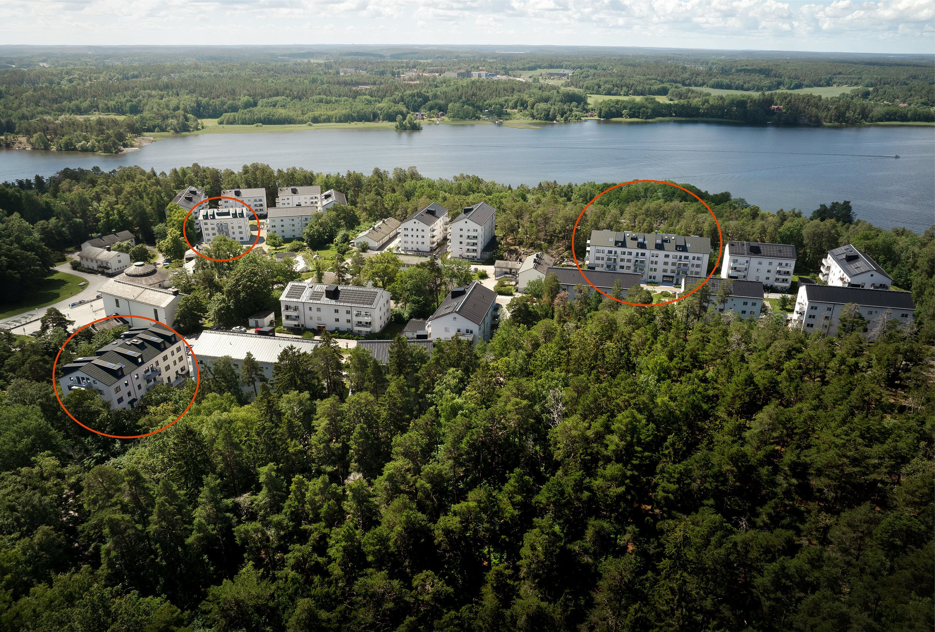 Flygbild där flera vita flerfamiljsbyggnader ligger i en grön miljö där vatten anas i bakgrund. Tre byggnader är markerad med orange ring.