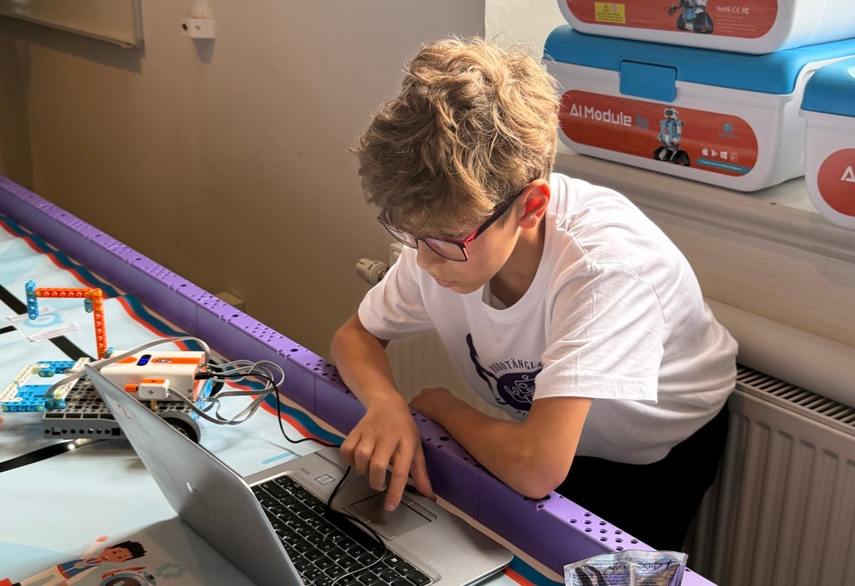 Pojke som arbetar vid en laptop på ett robotbord nära ett fönster, omgiven av färgglada robotlådor och elektronik.