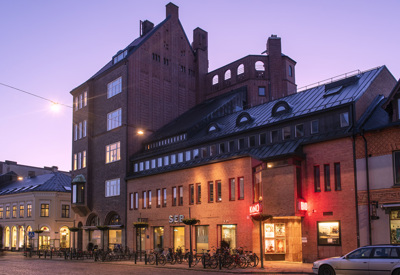 En tegelbyggnad i stadsmiljö fotograferad i skymningsljus. Byggnaden har en unik arkitektur med flera nivåer och tak i olika vinklar, samt en bioskylt med texten 'KINO' belyst i rött. Framför byggnaden finns cykelställ med många cyklar, och till vänster syns en rad äldre hus med välvda fönster och takkupor.
