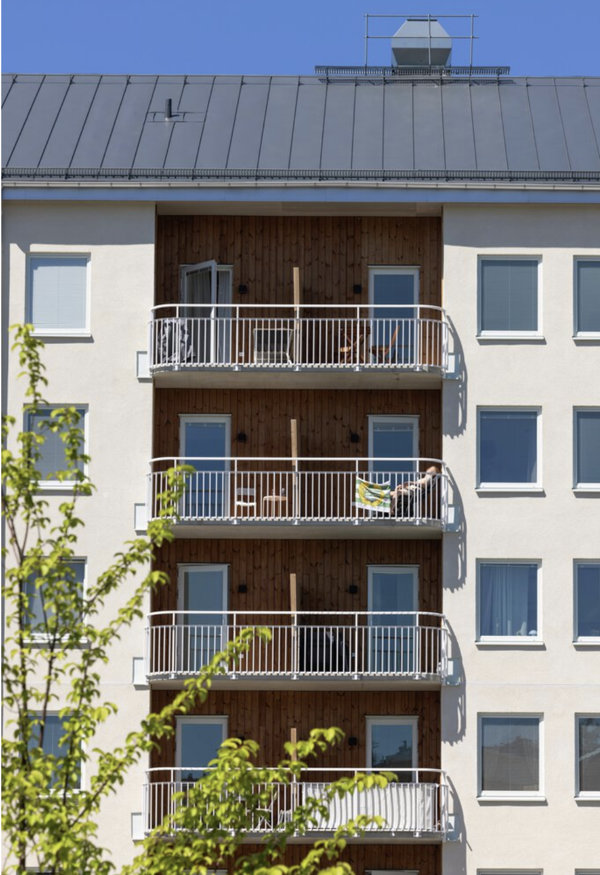 Fasad av ett modernt flerbostadshus med vita väggar och en mittsektion i trä, där flera balkonger med vita räcken ligger ovanpå varandra; på balkongerna syns utemöbler och en person som solar, och i förgrunden skymtar gröna träd mot en klarblå himmel.
