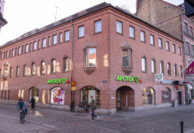 Tegelfasad på ett hörnhus i stadsmiljö med två apoteksskyltar i grönt som visar 'Apotek Hjärtat'. Byggnaden har stora skyltfönster i bottenplan och många små fönster på de övre våningarna. Flera personer promenerar och cyklar på den kullerstensbelagda gatan utanför, och en butiksskylt med ett hjärta syns till höger.
