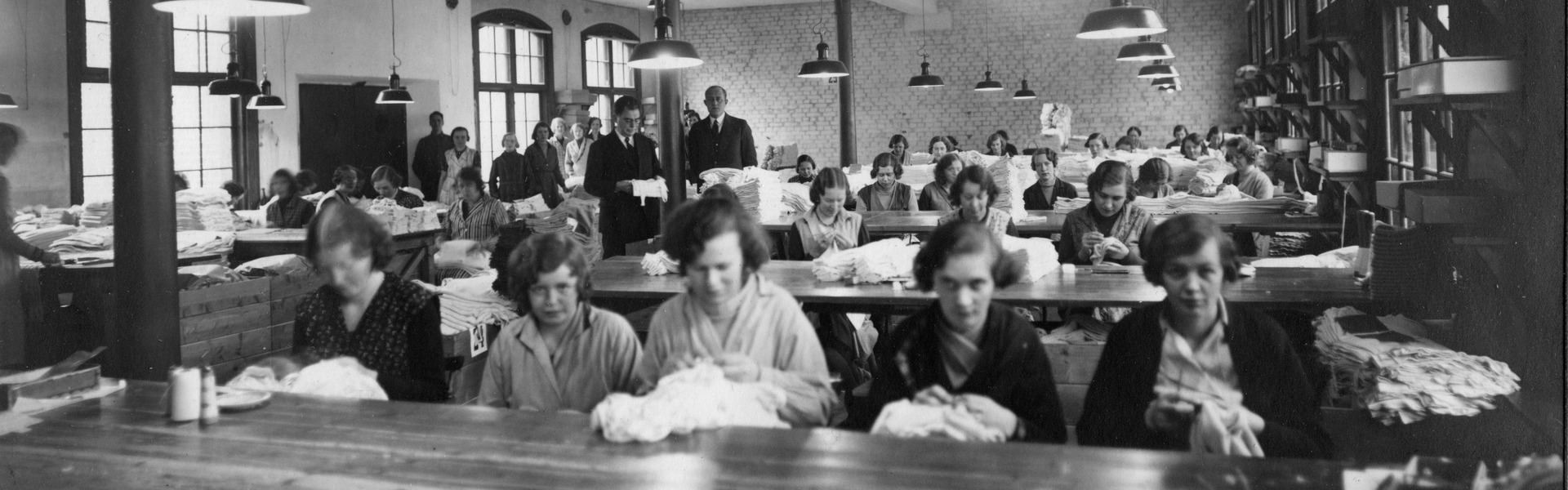 En svartvit historisk bild från en stor arbetslokal där flera personer sitter vid långa bord och arbetar med textilier. I rummet syns taklampor, stora fönster och högar av tyg på borden.