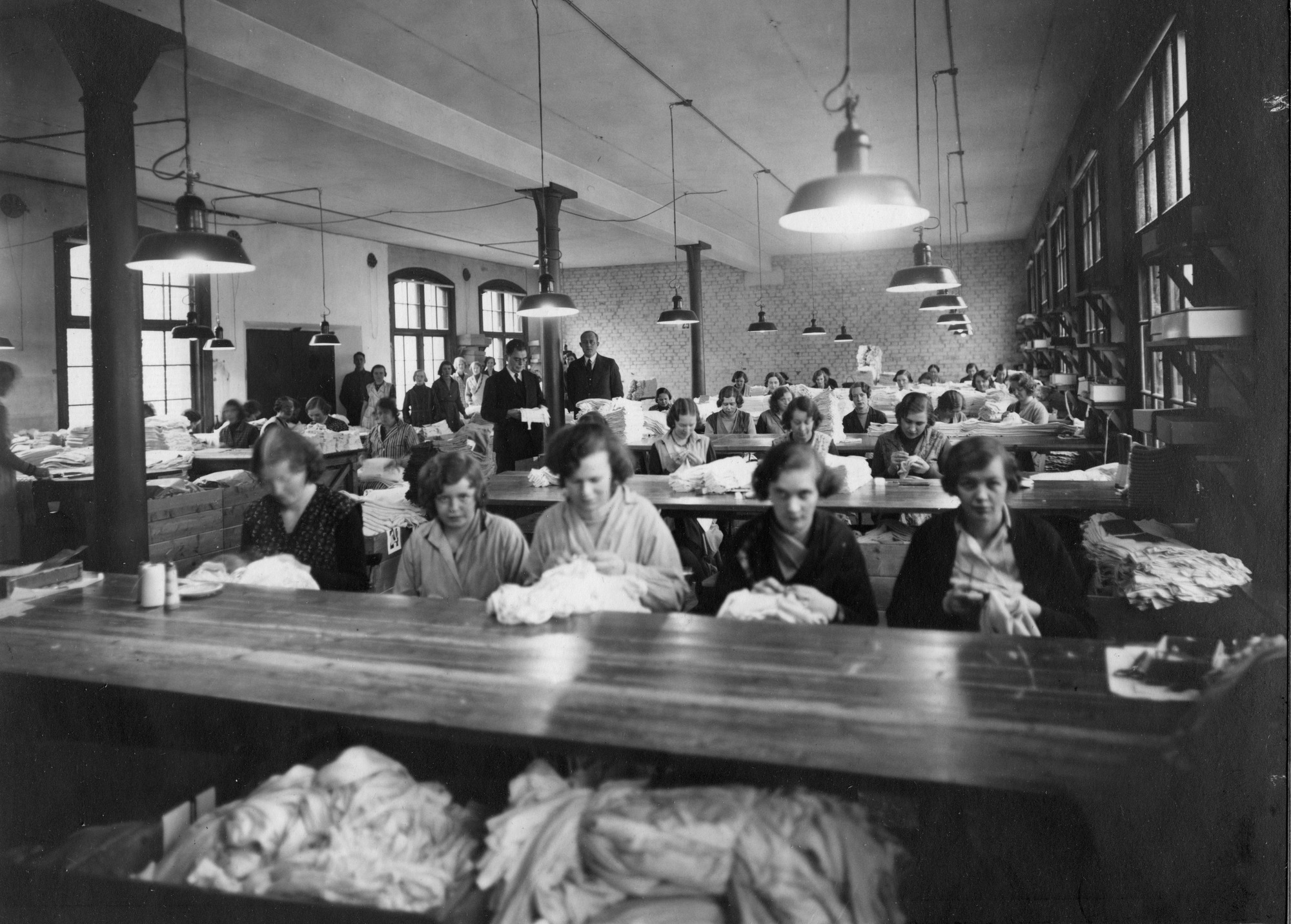En svartvit historisk bild från en stor arbetslokal där flera personer sitter vid långa bord och arbetar med textilier. I rummet syns taklampor, stora fönster och högar av tyg på borden.