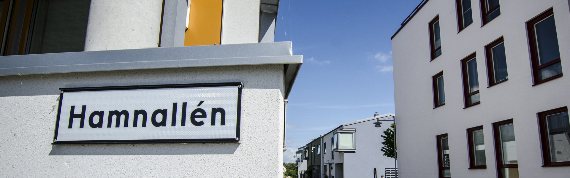 En gata med namnet "Hamnallén" på en skylt i förgrunden. Till vänster syns en byggnad med vita väggar och grå detaljer. Till höger sträcker sig en rad av moderna, vita flerfamiljshus med röda fönsterkarmar längs en asfalterad väg under en klarblå himmel.