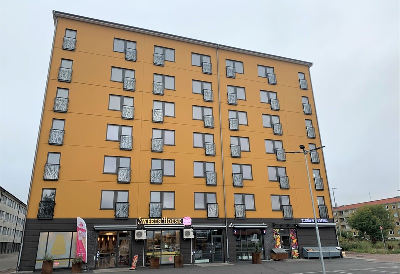 En flerfamiljsbyggnad med en senapsgul fasad och flera våningar. På bottenvåningen finns butiker, inklusive en med skylten "Sweet House". Himlen är molnig och omgivningen är urban.