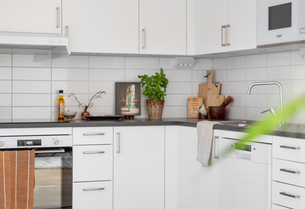 Vit, modern köksinredning i vinkel med släta skåpsluckor, mörk bänkskiva och vitt kakel. På bänken står kryddor, skärbrädor och en grön krukväxt som skapar en ombonad känsla. I nederkant syns grått klinkergolv och en del av en stoppad stol.