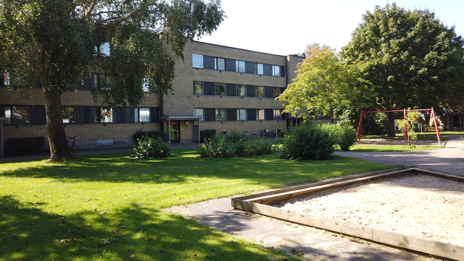 Grön innergård i solsken med gräsmatta, träd, sandlåda och gungställning framför ett trevånings flerbostadshus i tegel.