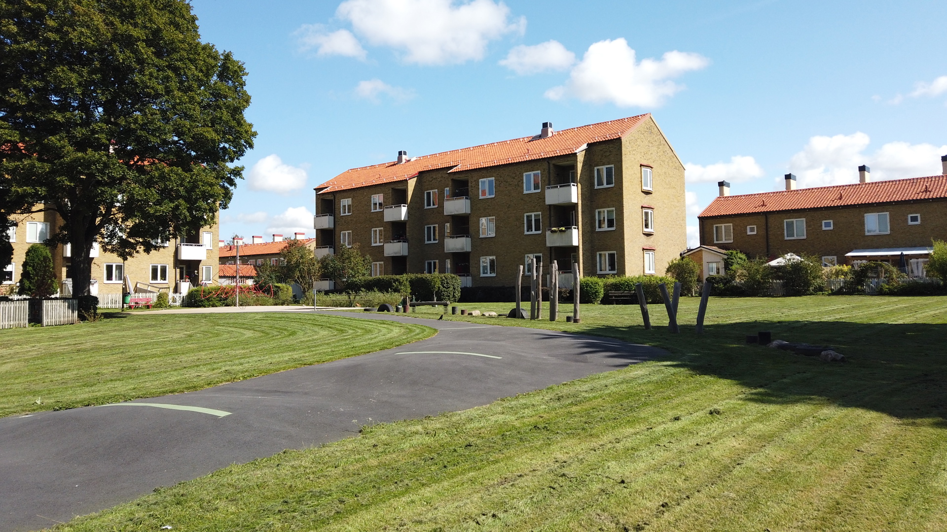 Grön innergård i solsken med träd, buskar, blommande planteringar, gångstig och en sandlåda längst fram, omgiven av flerbostadshus i tegel.