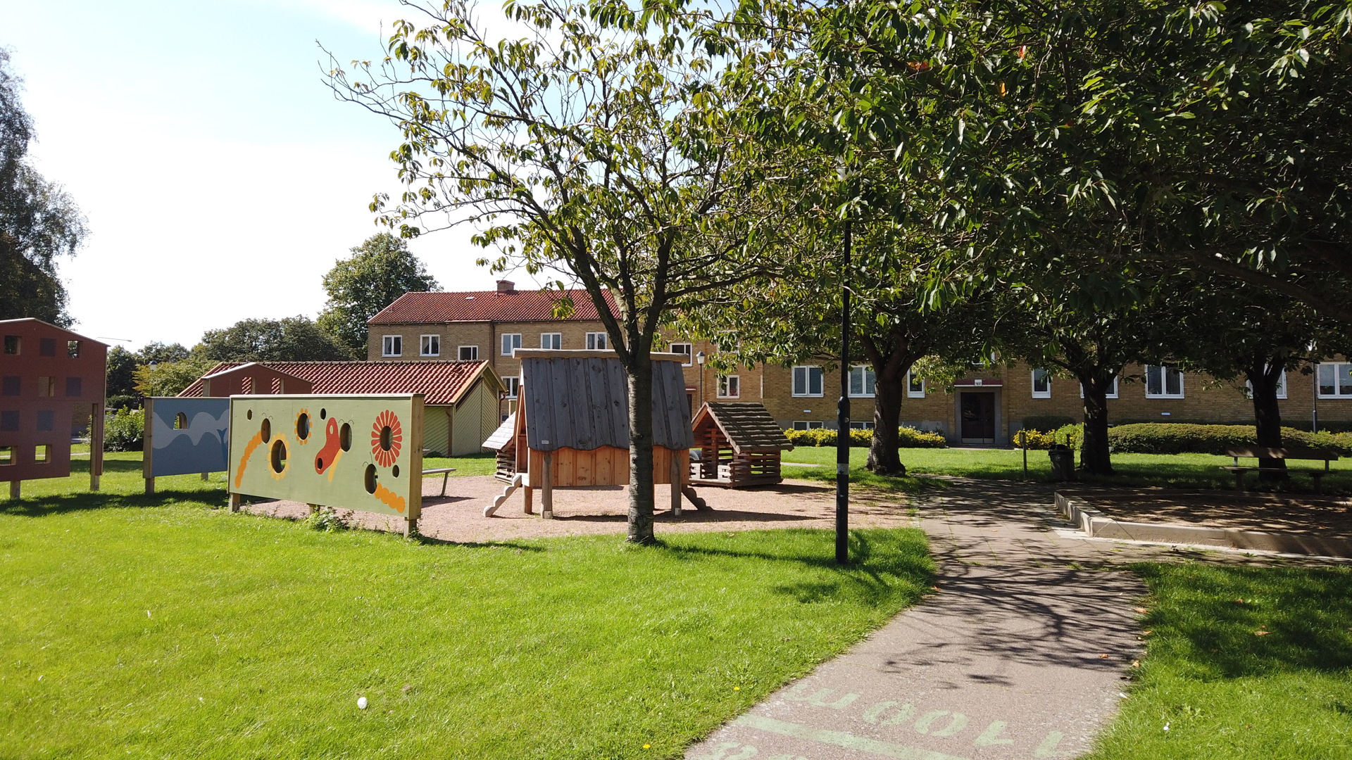 Solbelyst innergård med grön gräsmatta, gångväg, lekplats med sandlåda, litet lekhus och färgglad målad skärm, omgiven av trevånings flerbostadshus i tegel.