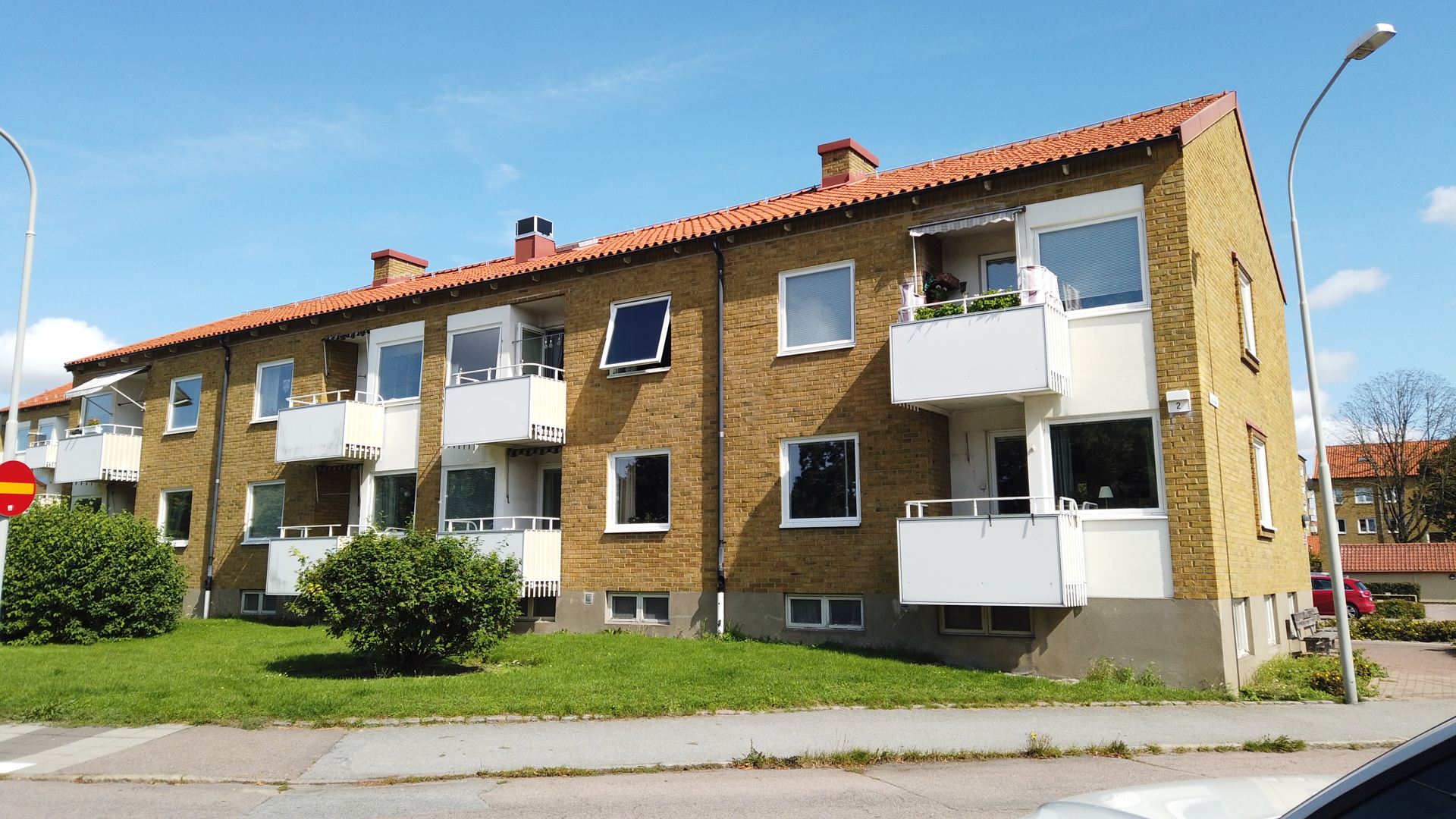 Trevånings flerbostadshus i gult tegel med vita balkonger och rött tegeltak, sett från gatan med gräsmatta och gångbana i förgrunden.