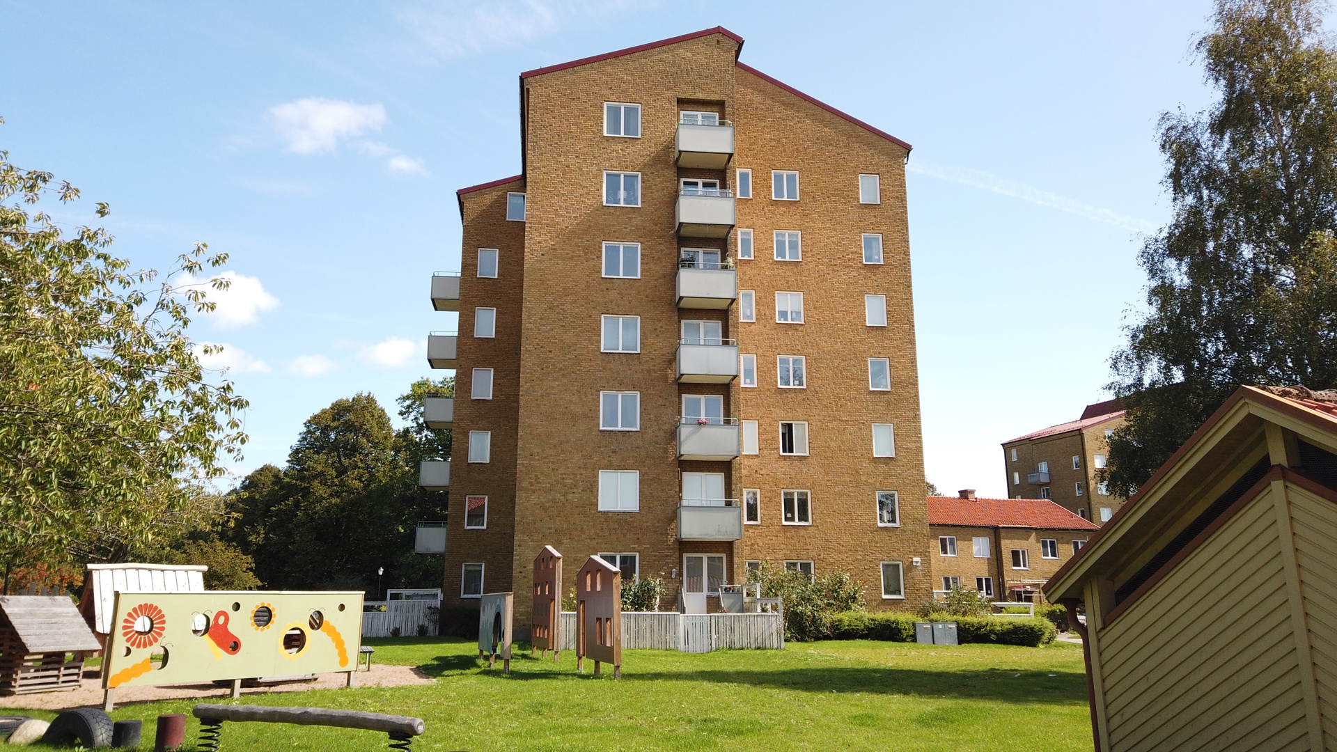 Sexvånings flerbostadshus i gult tegel med balkonger mot en solbelyst gräsmatta med lekplatsutrustning och färgglad målad skärm i förgrunden.