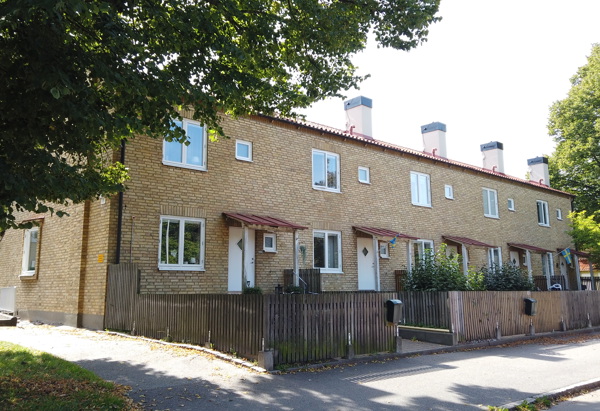 Rad med tvåvånings radhus i gult tegel med små entrétak och trästaket längs en lugn gata, delvis skuggade av stora träd.