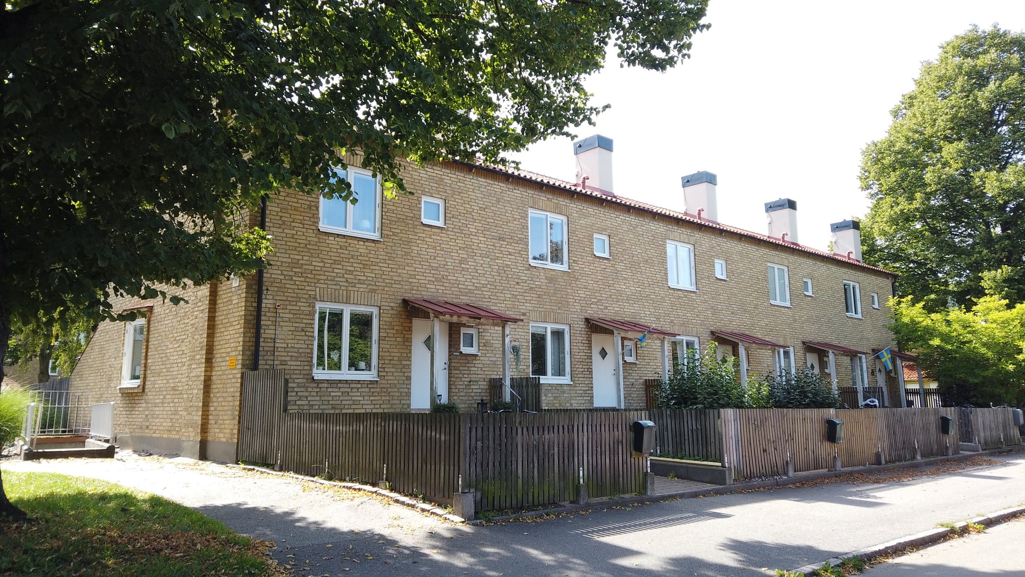 Rad med tvåvånings radhus i gult tegel med små entrétak och trästaket längs en lugn gata, delvis skuggade av stora träd.