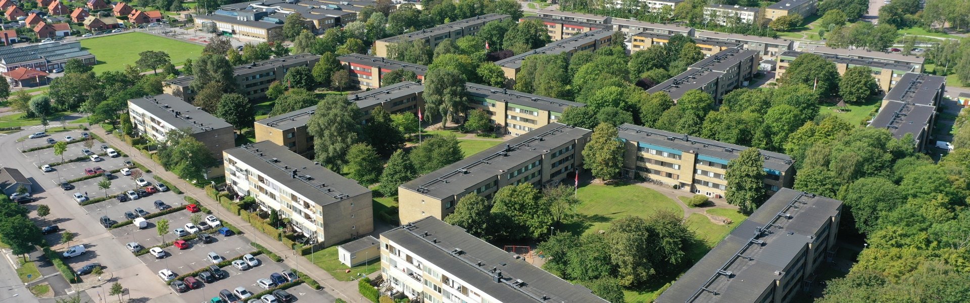 Flygfoto över ett bostadsområde med flerbostadshus, parkeringsplatser och gröna ytor. I bakgrunden syns vatten och bebyggelse.