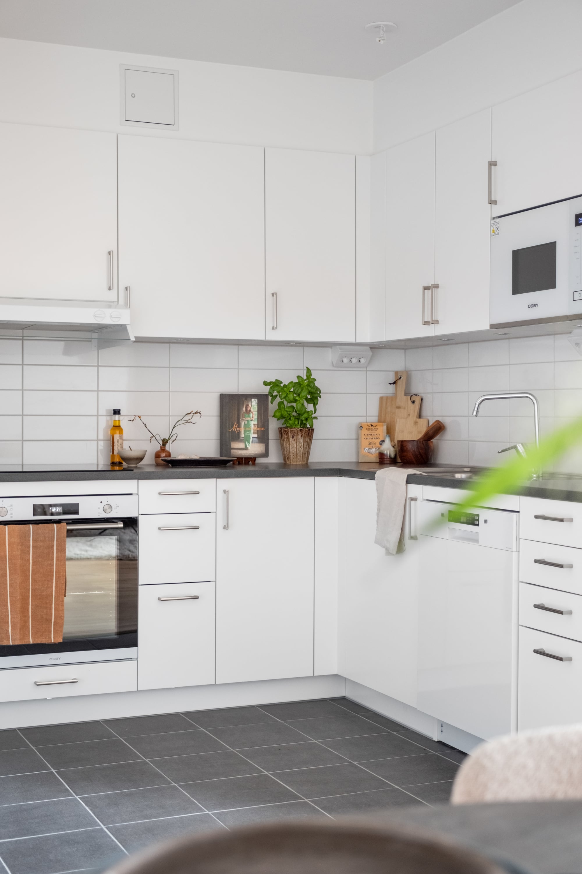 Vit, modern köksinredning i vinkel med släta skåpsluckor, mörk bänkskiva och vitt kakel. På bänken står kryddor, skärbrädor och en grön krukväxt som skapar en ombonad känsla. I nederkant syns grått klinkergolv och en del av en stoppad stol.