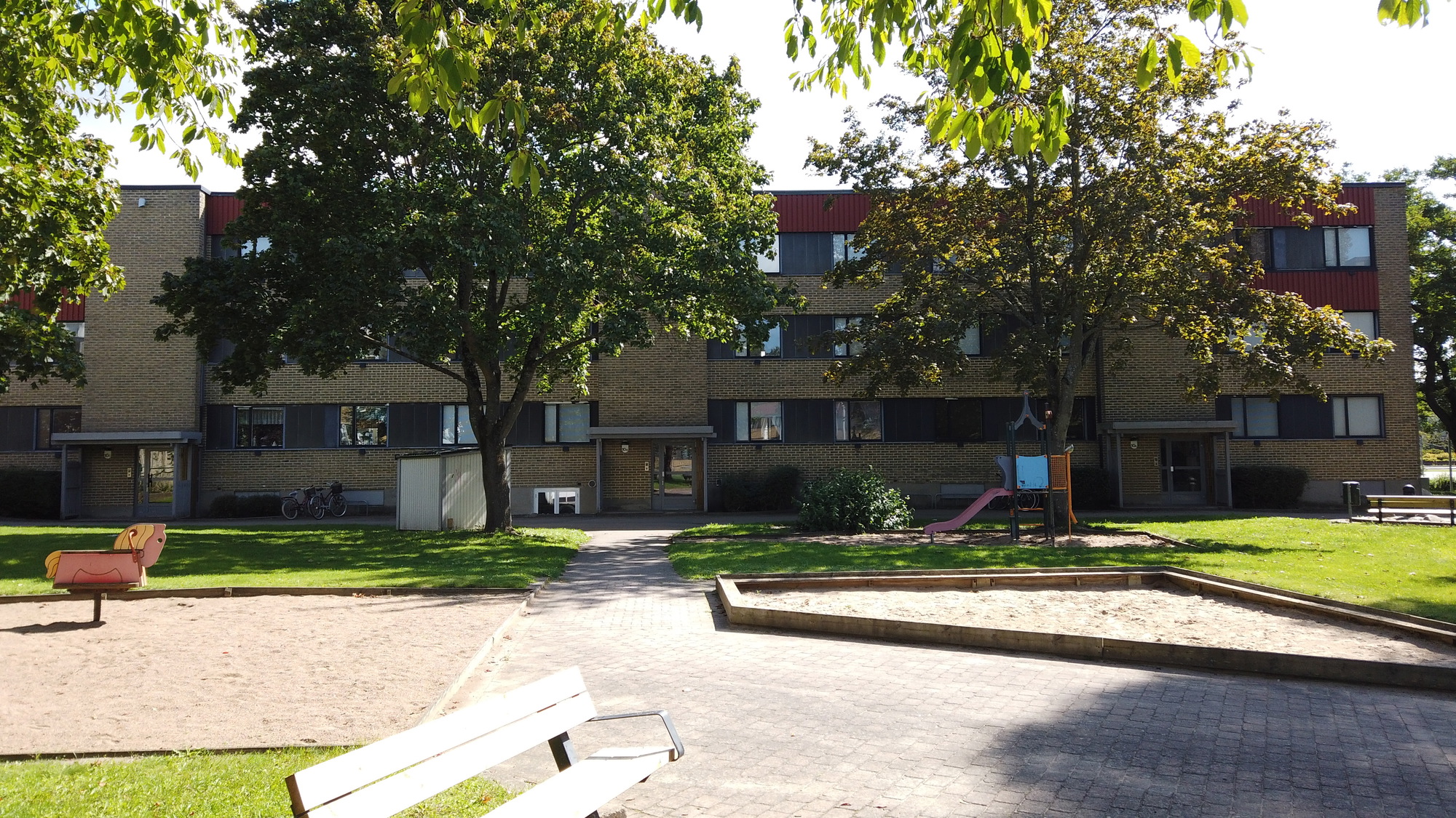 En solig innergård med grön gräsmatta, träd, sandlåda, lekplatsutrustning och parkbänkar framför ett trevånings flerbostadshus i tegel.