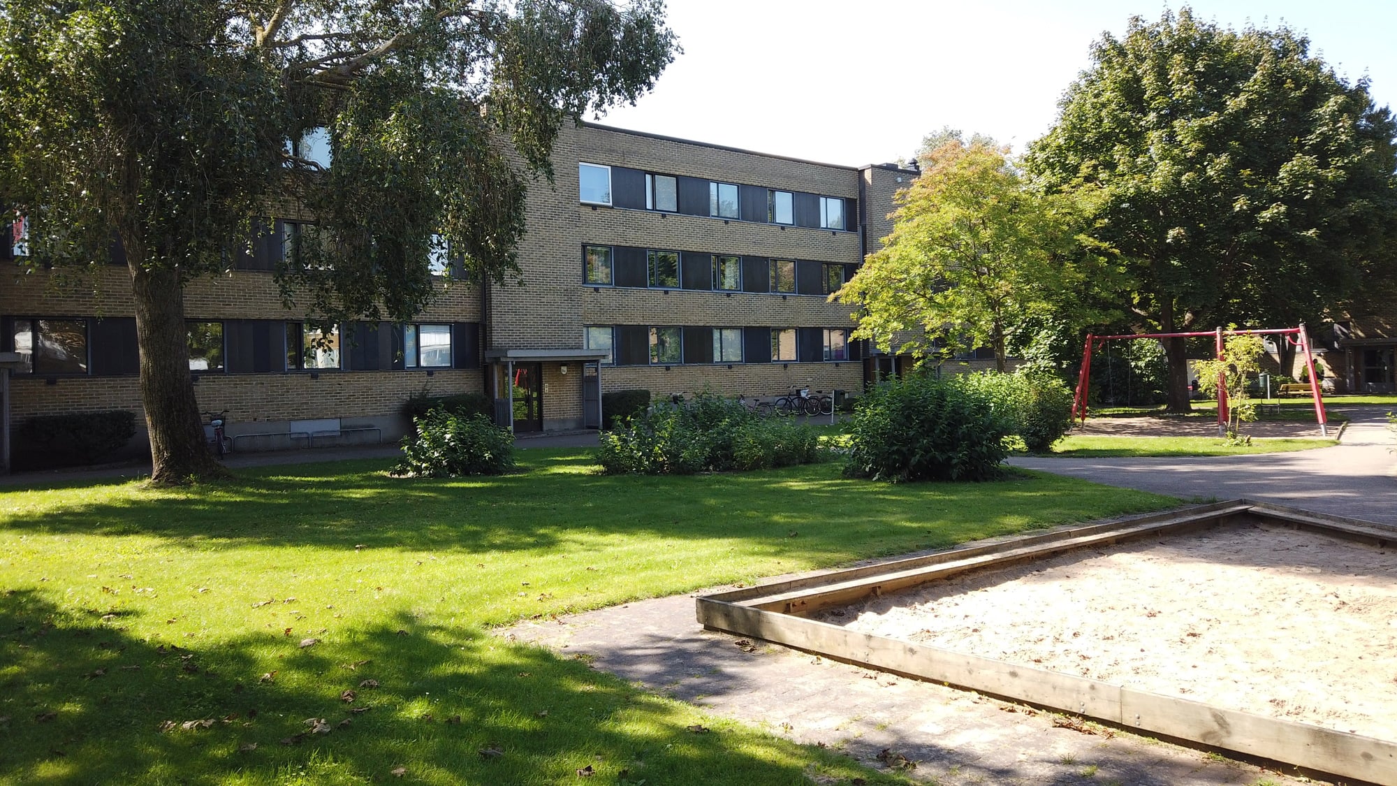 Grön innergård i solsken med gräsmatta, träd, sandlåda och gungställning framför ett trevånings flerbostadshus i tegel.