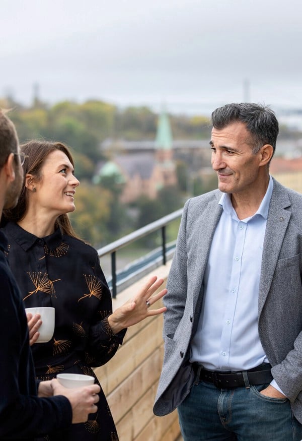 Tre personer står på en takterass och samtalar, stadsvy i bakgrunden
