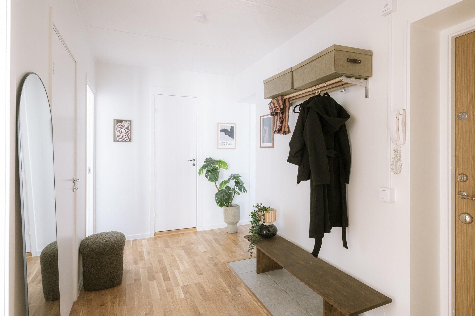 Ljus och rymlig hall med ljust trägolv, vitmålade väggar, en träbänk med växt och ljuslykta, ytterkläder hängande på en hatthylla samt spegel, sittpuff och gröna krukväxter som inredning.