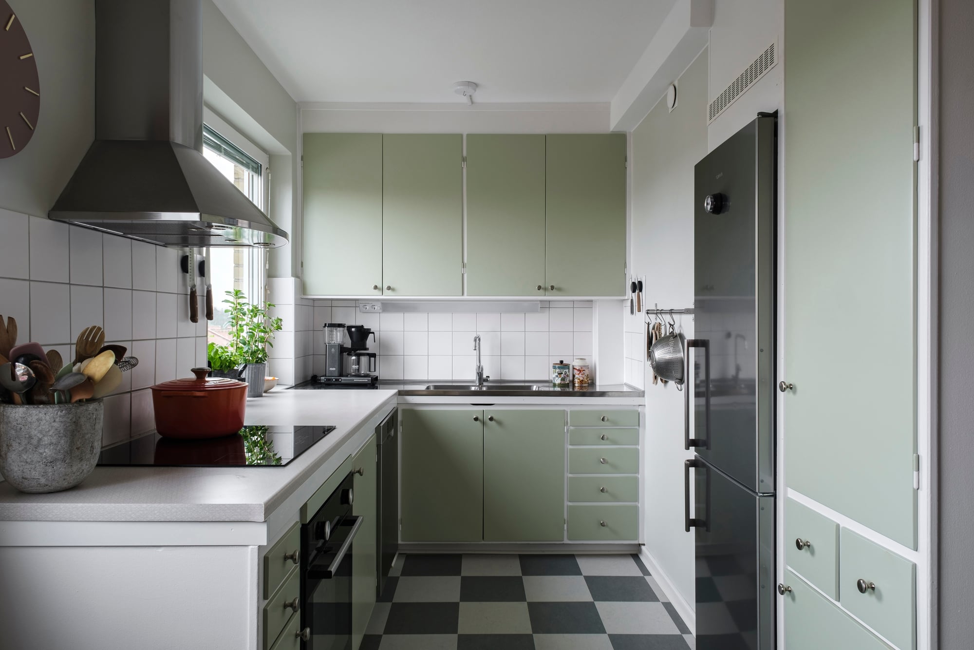 En modern kök med ljusgröna skåpluckor och en svartvit rutigt golv. Köket har en rostfri diskbänk med en kran, en spis med ugn och en fläkt ovanför. Till vänster står en kruka med köksredskap och en röd kastrull på spisen. Ett fönster släpper in naturligt ljus och det finns en kaffebryggare på bänken.