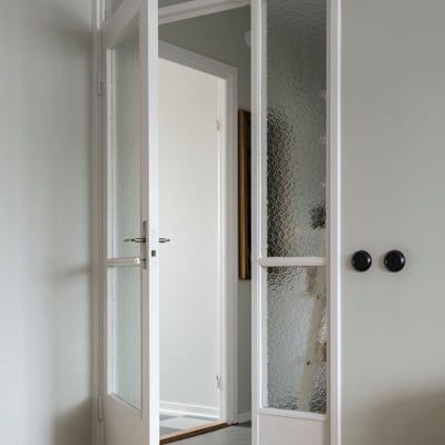 En vit dörr med glaspartier står på glänt och leder till en ljus hall. Dörren har svarta, runda handtag och texturerat glas för avskildhet. Golvet är av ljust trä i ett fiskbensmönster.