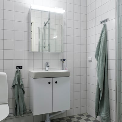 Ett badrum med vita kaklade väggar. Det finns ett handfat med ett spegelskåp ovanför, belyst av en lampa. Till vänster syns en del av en toalettstol och två gröna handdukar hänger på krokar. Golvet är rutigt i grå nyanser.