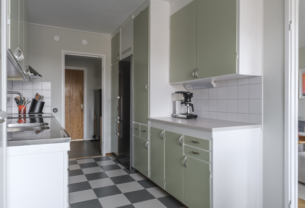 Ett kök med ljusgröna skåpluckor och ett svartvitt rutigt golv. På bänken står en kaffebryggare och köksredskap i en behållare. Till vänster syns en rostfri diskbänk och spis. En öppen dörr leder till ett angränsande rum.
