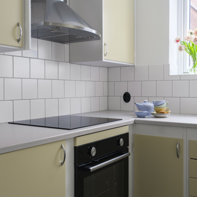 Ett kök med ljusgula skåpluckor och vita kaklade väggar. En svart spishäll och ugn är placerade under en rostfri köksfläkt. På bänken vid fönstret står staplade koppar och en vas med blommor.