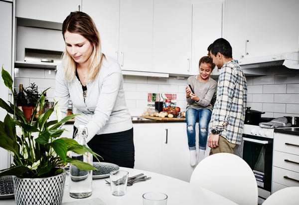 En kvinna dukar ett bord i ett modernt kök. I bakgrunden sitter en ung kvinna på köksbänken och visar något på sin mobil för en man som står bredvid. Köket är ljust med vita skåp och en växt på bordet.