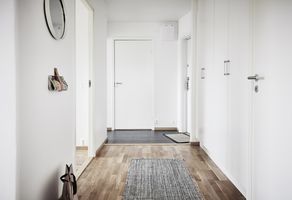 En ljus hall med vita väggar och trägolv. En grå matta ligger på golvet, och det finns en rund spegel på väggen. Dörrar leder till andra rum, och en krok med nycklar hänger på väggen.