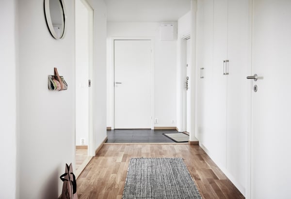 En ljus hall med vita väggar och trägolv. En grå matta ligger på golvet, och det finns en rund spegel på väggen. Dörrar leder till andra rum, och en krok med nycklar hänger på väggen.