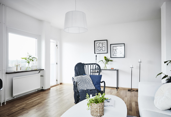 Ett ljust vardagsrum med trägolv och vita väggar. En soffa och en fåtölj med blå kuddar står vid ett ovalt soffbord med en grön växt. På väggen hänger två tavlor, och en golvlampa står i hörnet. Ett fönster släpper in naturligt ljus.