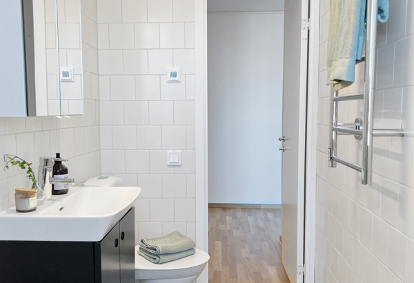 Ett ljust badrum med vita kakelväggar och grått klinkergolv. En svart kommod med handfat och spegel ovanför. På handfatet finns en tvålflaska och en liten växt. En handdukshängare med handdukar är monterad på väggen, och en dörr leder ut till ett rum med trägolv.